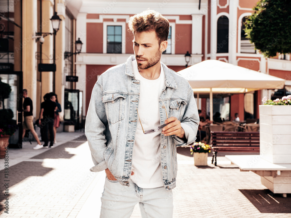 Fototapeta premium Portrait of handsome confident stylish hipster lambersexual model. Sexy man dressed in jeans jacket. Fashion male posing in the street in sunglasses in Europe city. Outdoors at sunset