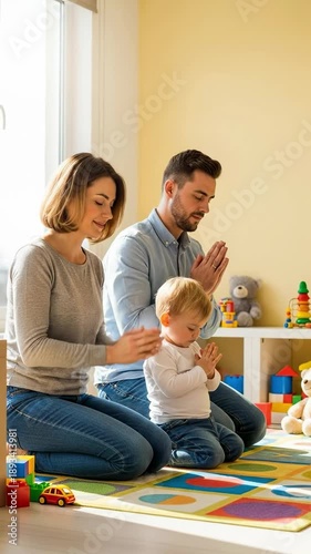Young Caucasian family with a small toddler boy kneeling and praying together in a bright playroom with toys, teaching faith and spirituality at home.