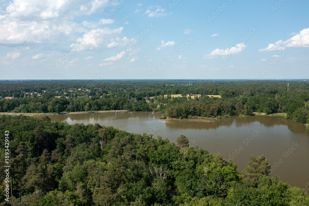Fototapeta premium Expansive aerial landscape featuring a calm forest lake bordered by dense green woodland, with a rural village and open fields visible on the horizon under a bright blue sky with scattered clouds near