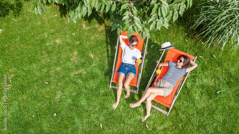 Fototapeta premium Young girls relax in summer garden in sunbed deckchairs on grass, women friends have fun outdoors in green park on weekend, aerial drone view from above