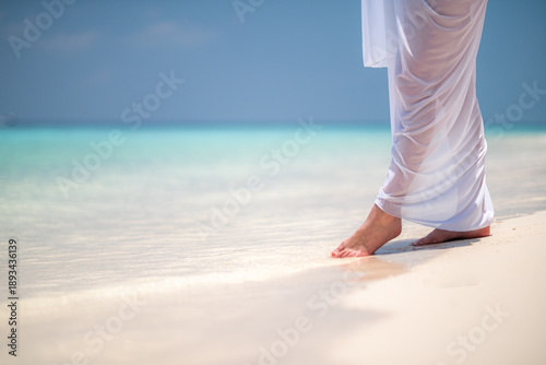 Obraz na plátně woman with bare legs in white sarong stands in calm blue sea, gently dipping her feet, evoking tranquility and peace, serenity, relaxation, escape