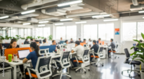 Blurred busy modern openplan office environment with many employees working at desks with computers, bright lighting, and large windows creating a dynamic corporate workspace atmosphere