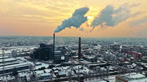 Aerial winter view of industrial power plant with smokestacks releasing steam over snowy city at sunrise, highlighting energy production, pollution, and urban infrastructure.