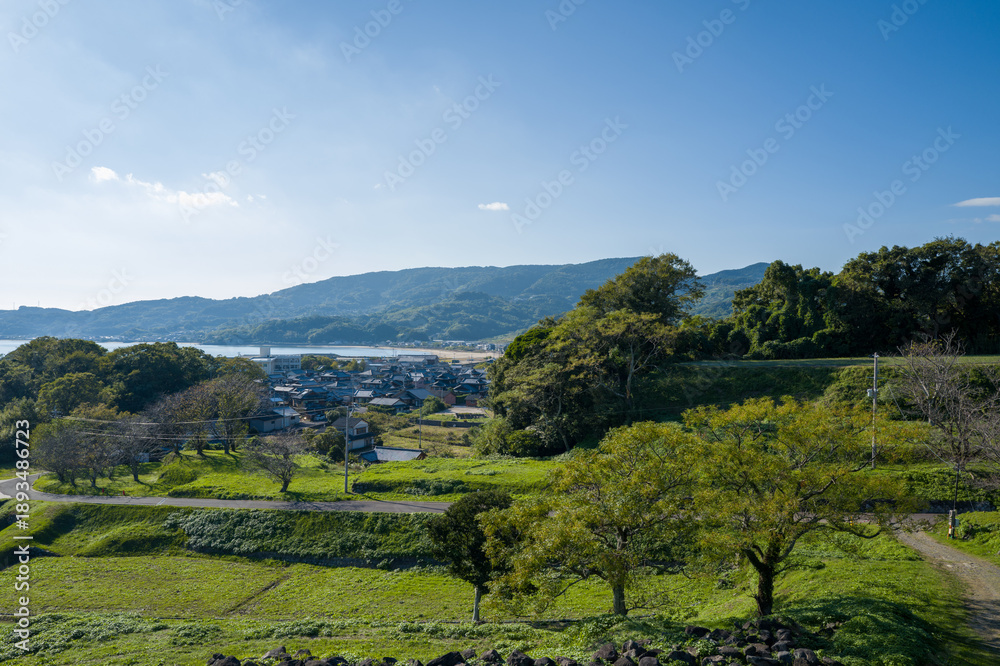 Obraz premium A peaceful rural village nestles among lush green hills and scattered trees under a clear blue sky near Hara, with distant mountains and water visible in the background. Crisp sunlight highlights the
