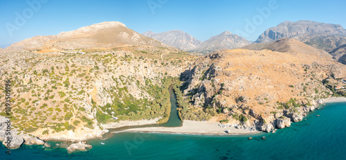 Sweeping aerial view of the Preveli river flowing through a lush palm-lined gorge to meet the sandy beach and turquoise Mediterranean Sea, surrounded by arid mountains and rugged cliffs under a clear