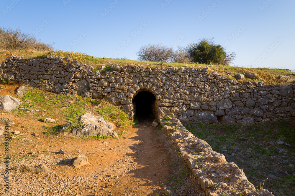 Fototapeta premium A weathered stone tunnel leads into the hillside at Palamidi Fortress in Nafplio, Greece. Sunlight highlights the rough masonry, dry earth, and sparse vegetation, creating a sense of mystery and