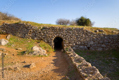 A weathered stone tunnel leads into the hillside at Palamidi Fortress in Nafplio, Greece. Sunlight highlights the rough masonry, dry earth, and sparse vegetation, creating a sense of mystery and