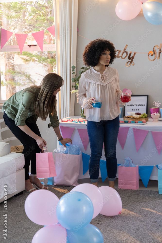 Naklejka premium Diverse female friends preparing drinks near gift bags, balloons in living room