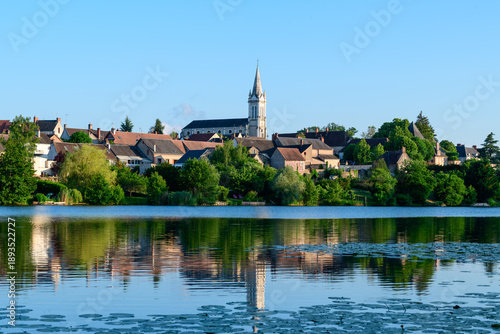 Quadro em tela Warm sunlight bathes the rooftops and tall church spire of Dampierre en Burly, their forms mirrored in the still waters of a pond bordered by lush summer foliage