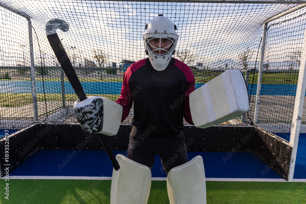 Fototapeta premium Field hockey goalkeeper is standing before goal cage on turf with stick wearing protective gear