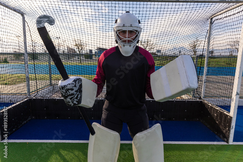 Field hockey goalkeeper is standing before goal cage on turf with stick wearing protective gear
