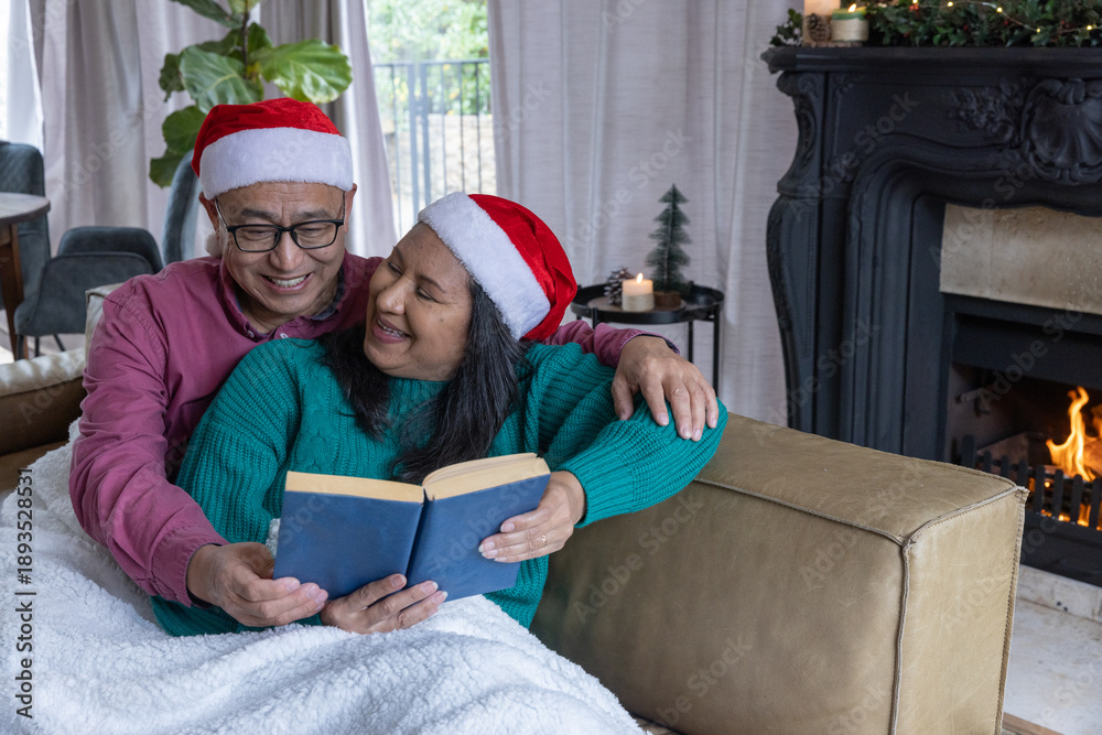 Naklejka premium Diverse couple in red santa hats sitting on leather couch reading blue book in living room
