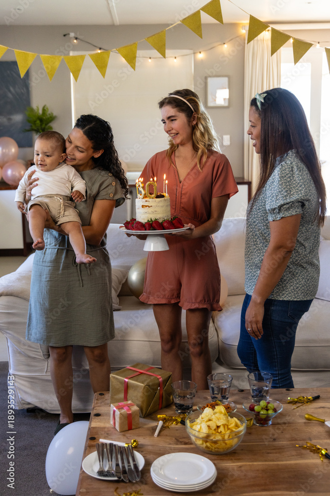 Naklejka premium Diverse female friends celebrating in living room holding cake with lit candles, cradling baby