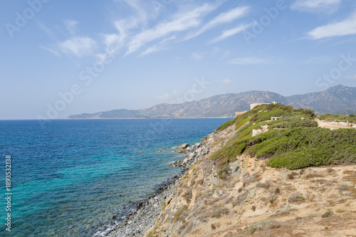 Wallpaper Mural Sunlit rocky cliffs covered in green shrubs rise above clear turquoise waters at Cala Caterina, with distant mountains and a bright blue sky creating a serene Mediterranean landscape. Torontodigital.ca