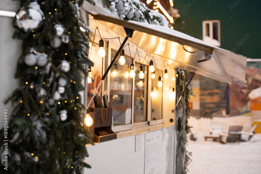 Fototapeta premium Cozy street cafe wagon at lively city square decorated with christmas lights and fir tree garland covered in snow. Bistro with new year's festive decorations on roof and window during winter season.