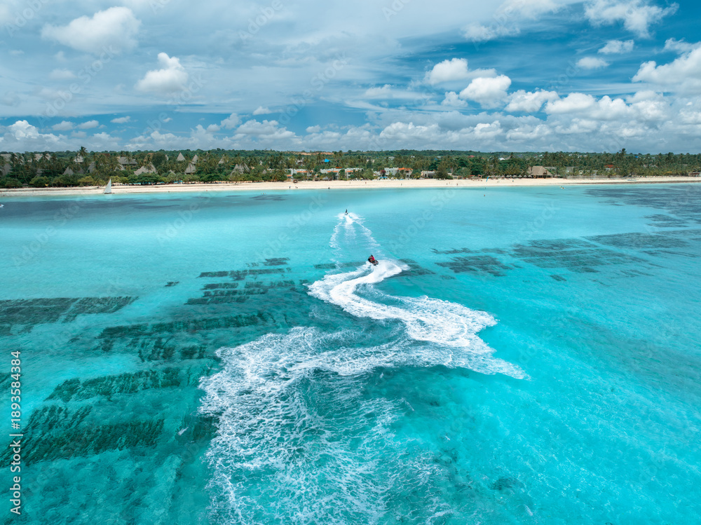 Fototapeta premium Aerial view of jet skis over shallow turquoise sea, sandy beach, palm trees in Zanzibar. Clear tropical water, watercraft trails, summer vacation, coastal travel and marine leisure activity. Top view