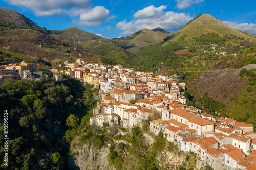 Aerial image of Tortora, Italy, featuring a cluster of stone houses with terracotta roofs perched on a steep cliff, surrounded by lush hills and rugged mountains beneath a bright sky with scattered