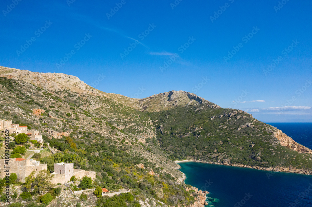 Obraz premium Aerial view of a tranquil bay framed by rugged hills and traditional stone buildings nestled among greenery on the Mani peninsula, Greece, under a vivid blue sky.