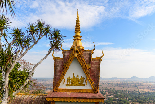 A detailed view of the ornate golden spire and gabled roof of Phnom Sampov temple rises above the rural landscape near Battambang, Cambodia. The scene features decorative Khmer motifs, tropical