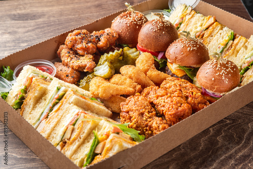 Foto A tempting assortment of party foods in a cardboard platter box, featuring mini cheeseburger sliders, crispy fried chicken pieces, and various club sandwiches on a wooden table