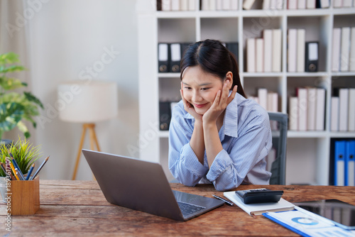 Tableau sur toile Asian woman smiling, working on her laptop at a wooden desk in an office or libr