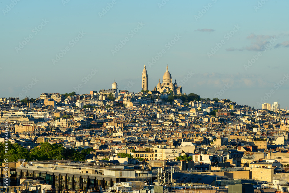 Obraz premium Sacre-Cur basilica rises above the densely packed rooftops of Montmartre, bathed in warm evening sunlight under a clear blue sky. The elevated view highlights the layered urban landscape and iconic