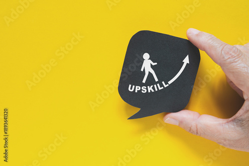 hand hold UPSKILL text , increasing arrow and man walking icon on real black speech bubble paper cut on yellow background