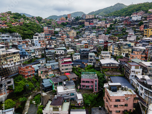 Aerial view of the mountain town of Jiufen in Taiwan, with dense hillside houses, winding streets, and dramatic terrain overlooking the coast, creating a layered and atmospheric townscape.

