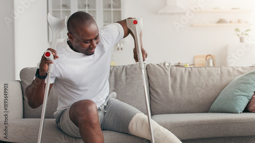 Schilderij op canvas A man with dark skin uses crutches while sitting on a couch in a living room