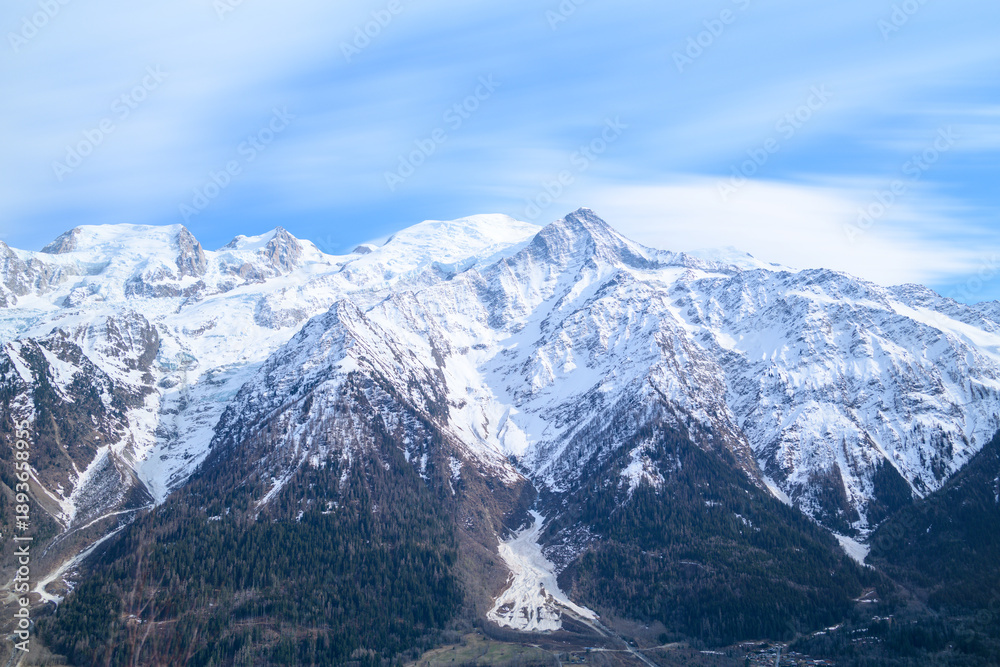 Obraz premium Snow-capped peaks of the Mont Blanc massif tower above dark pine forests and deep valleys, set against a sky streaked with wispy clouds.