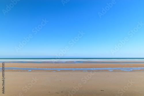 Foto Vast open sands and shallow tidal pools stretch across Omaha Beach beneath a cloudless, vibrant blue sky, creating a peaceful and spacious coastal scene
