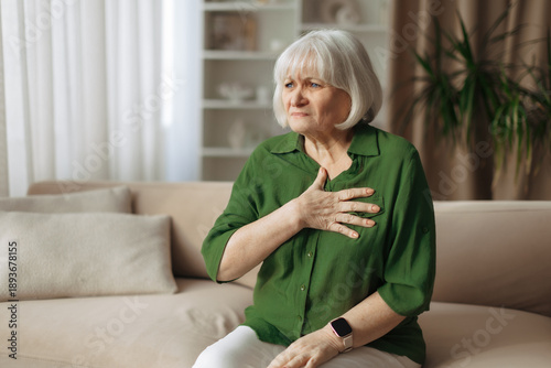Obraz na plátne Elderly caucasian woman holding chest while sitting on sofa at home, senior fema