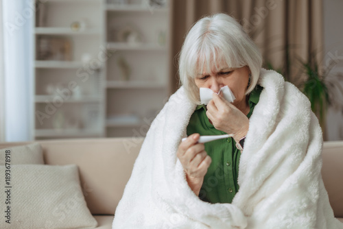 Fotografie Senior caucasian woman wrapped in blanket holding thermometer at home, elderly f