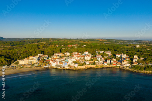 Εκτύπωση καμβά Colorful seaside houses line the shore of Arkoudi village, backed by lush green forest and open countryside under a clear blue sky
