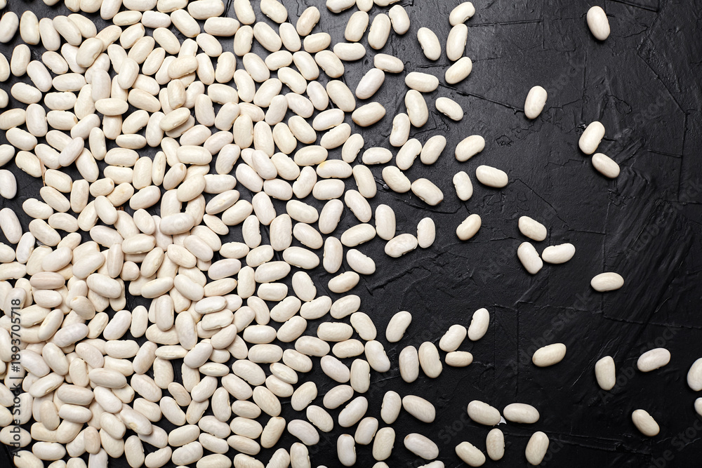 Fototapeta premium White beans scattered on a textured black surface, showcasing their smooth, oval shapes and natural color, ideal for culinary and health-related concepts with copy space