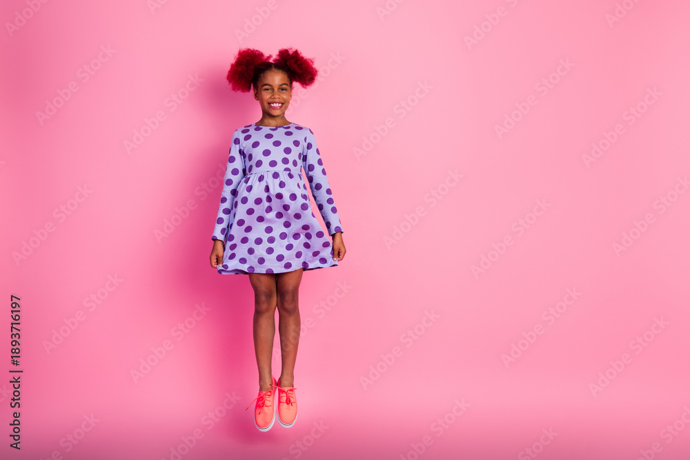 © deagreez - Young girl wearing purple polka dot dress jumping against pink background in a playful fashion lifestyle photo