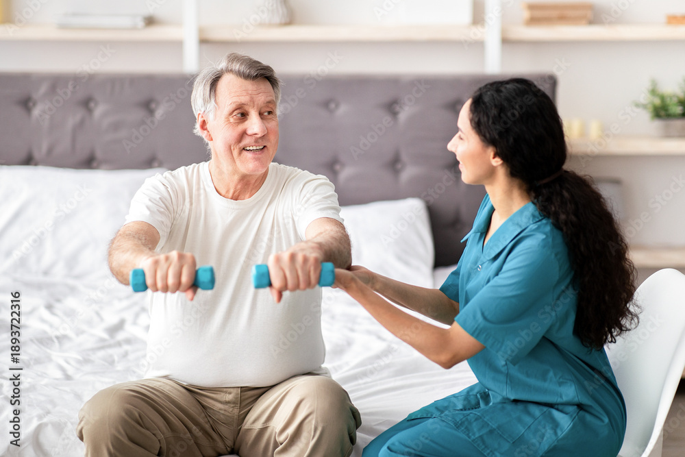 Obraz premium A senior man is sitting on a bed holding small weights as a woman provides encouragement and assistance. They are in a bright room designed for physical activity.