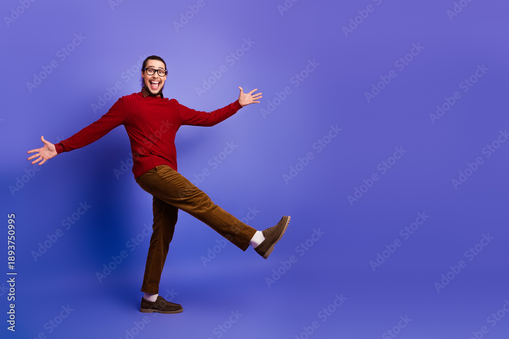 Fototapeta premium Happy young man in a red sweater balances on one leg against a violet backdrop for a lively fashion lifestyle stock photo