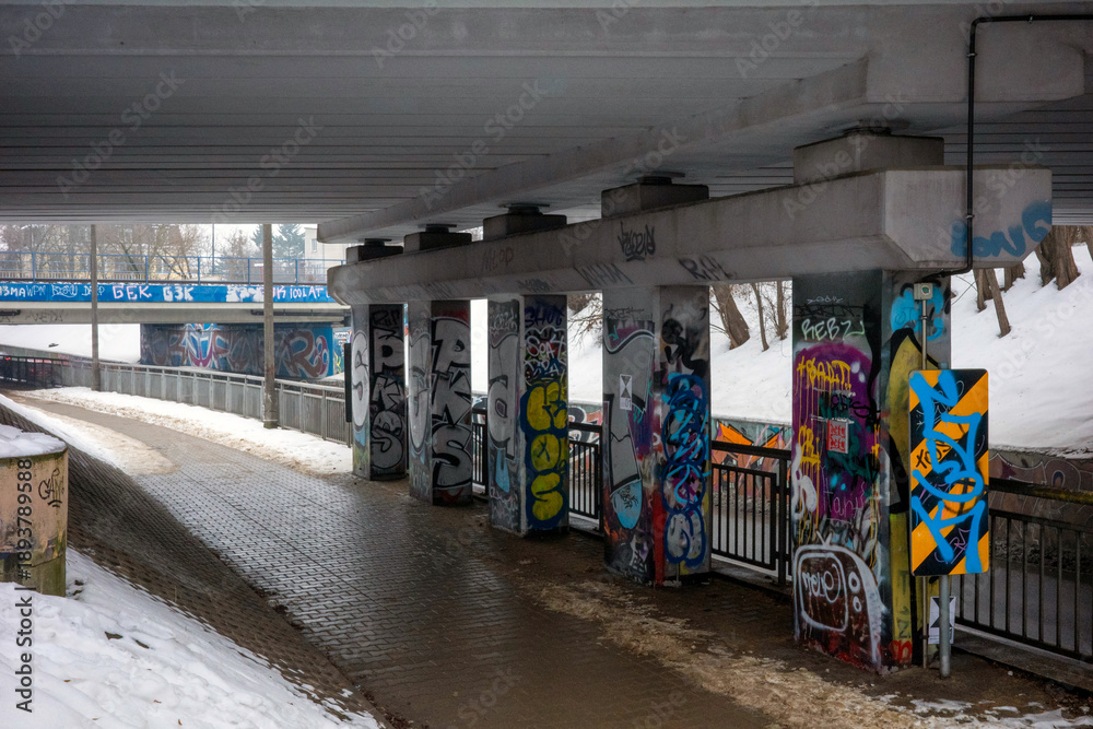 Fototapeta premium Warsaw, Masovian Voivodeship, Poland - January 27, 2026: A wide-angle shot of a pedestrian walkway beneath a concrete bridge structure heavily covered in vibrant graffiti art