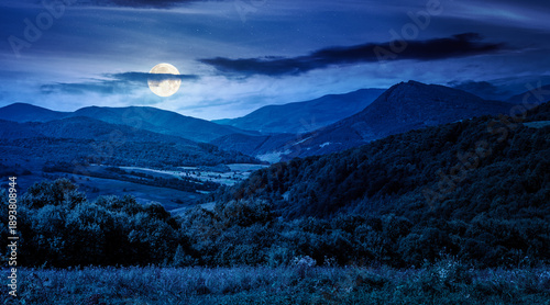 mountainous countryside scenery surrounded with forested rolling hills at night. rural area peaceful landscape with dark sky in full moon light. early autumn weather. mysterious fresh air environment
