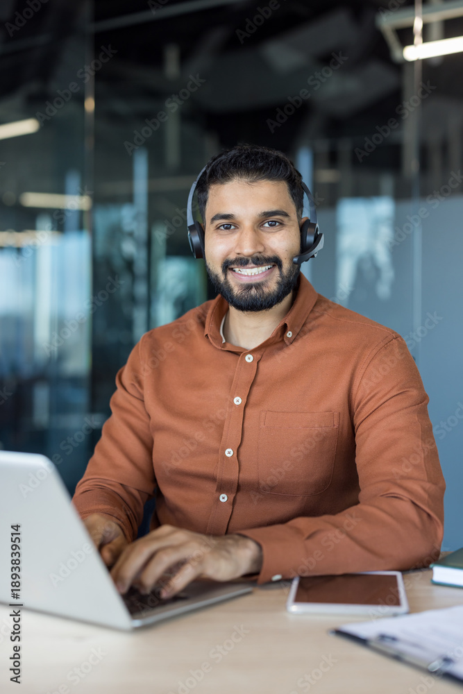 Obraz premium Smiling male call center agent wearing a headset, providing online assistance and technical support while typing on a laptop in a modern corporate office environment