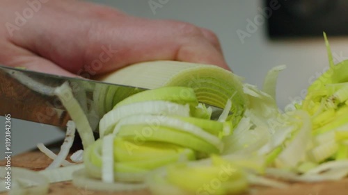 A hand cuts leeks on a wooden board in a kitchen. The green parts of the leeks are neatly arranged