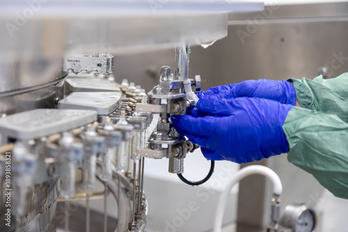 Hands encased in sterile blue gloves meticulously adjusting intricate components of an automated pharmaceutical manufacturing machine within a cleanroom environment.