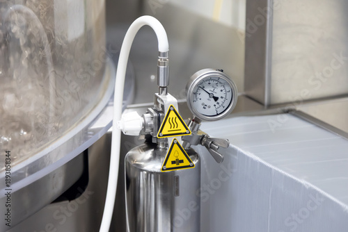 A close-up view of a stainless steel industrial pressure gauge and control valve with hot warning labels indicating operational heat.