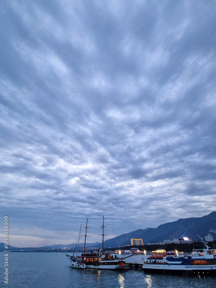 Fototapeta premium Docked sailboats and city lights at evening harbor with cloudy sky