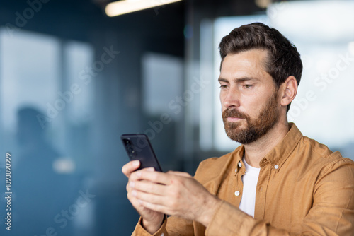 Young businessman concentrating on his smartphone for communication and technology tasks, managing business in a contemporary professional environment with a blue background