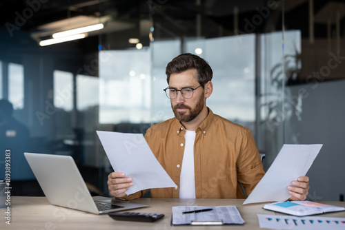Businessman focusing intently while analyzing paperwork and financial reports at his office desk, comparing information on documents with data displayed on a laptop screen