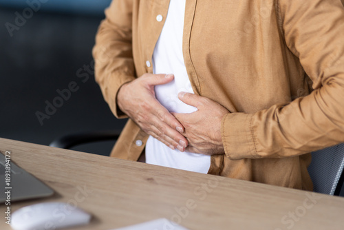 Man experiencing abdominal pain and discomfort, holding stomach while sitting at an office desk, illustrating health issues affecting productivity during workday