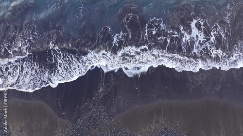 Top-down aerial drone footage showing a fixed overhead view of ocean waves breaking onto a dark shoreline. The shot emphasizes natural foam patterns, water movement.