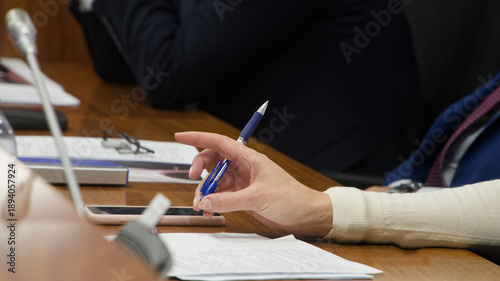 Female official sitting at wooden desk with colleagues during political summit. Hand with pen ready to sign contract or take notes near microphone system and eyeglasses. Photo
